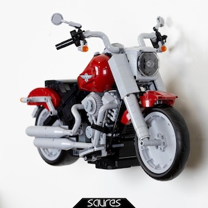 Op de afbeelding: Een rood en wit LEGO-motorfietsmodel met een zwarte motor en chromen uitlaatpijpen. De motorfiets is aan een muur gemonteerd.