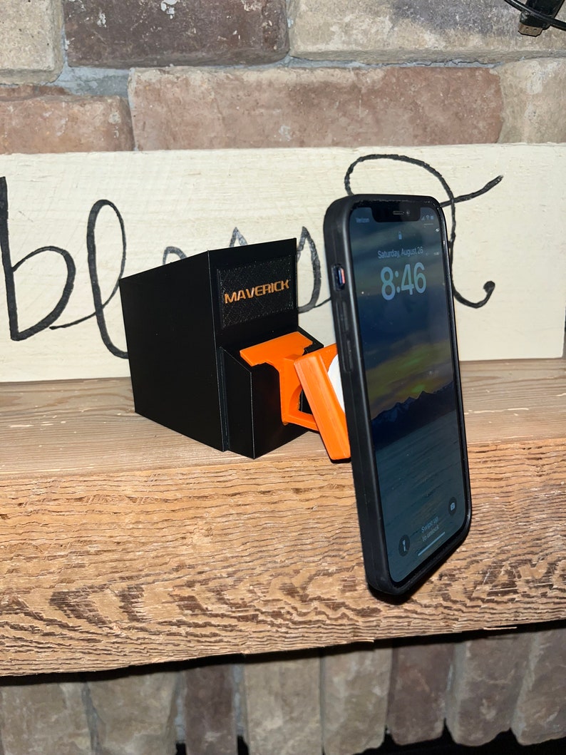 Puede incluir: Un soporte de tel&eacute;fono negro con la palabra "Maverick" impresa en &eacute;l. El soporte sostiene un tel&eacute;fono inteligente negro con una funda naranja brillante. La pantalla del tel&eacute;fono muestra la hora 8:46.