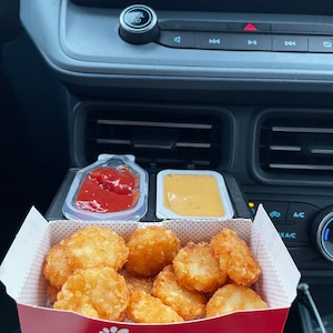 May include: A red Chick-fil-A box filled with golden brown hash browns. Two small containers with red and yellow sauces are visible in the background.
