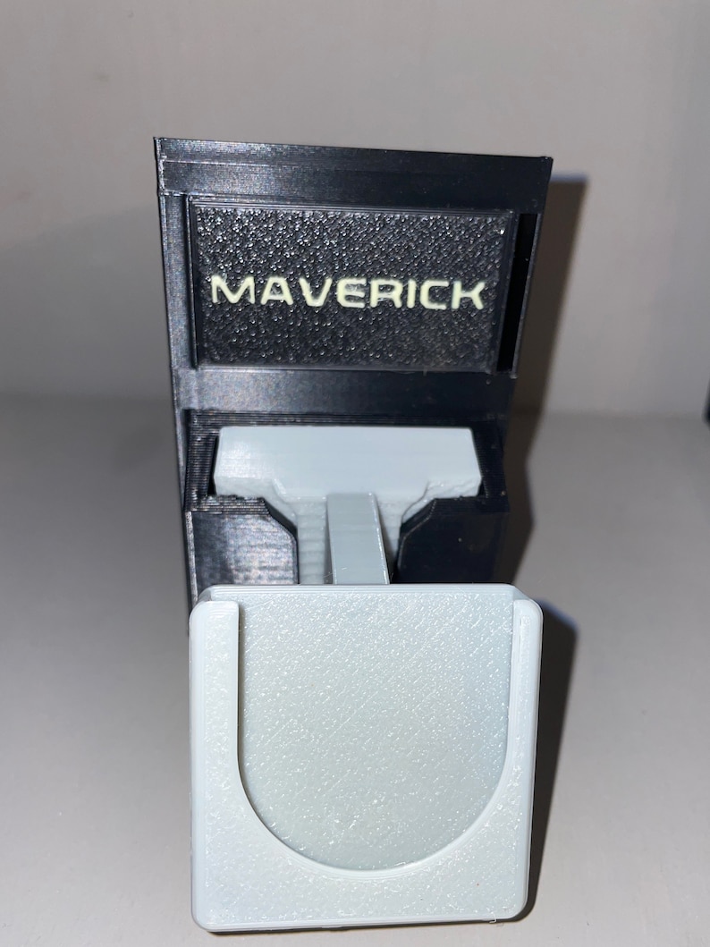 Puede incluir: Un objeto impreso en 3D en blanco y negro con la palabra "Maverick" impresa en la parte delantera. El objeto tiene una abertura en forma de U en la parte inferior.