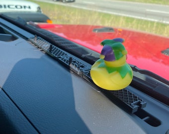 Puede incluir: Un pato de goma amarillo con un gorro de bufón verde y morado se sienta en un accesorio de tablero negro en un coche. El pato está mirando hacia el lado derecho de la imagen.