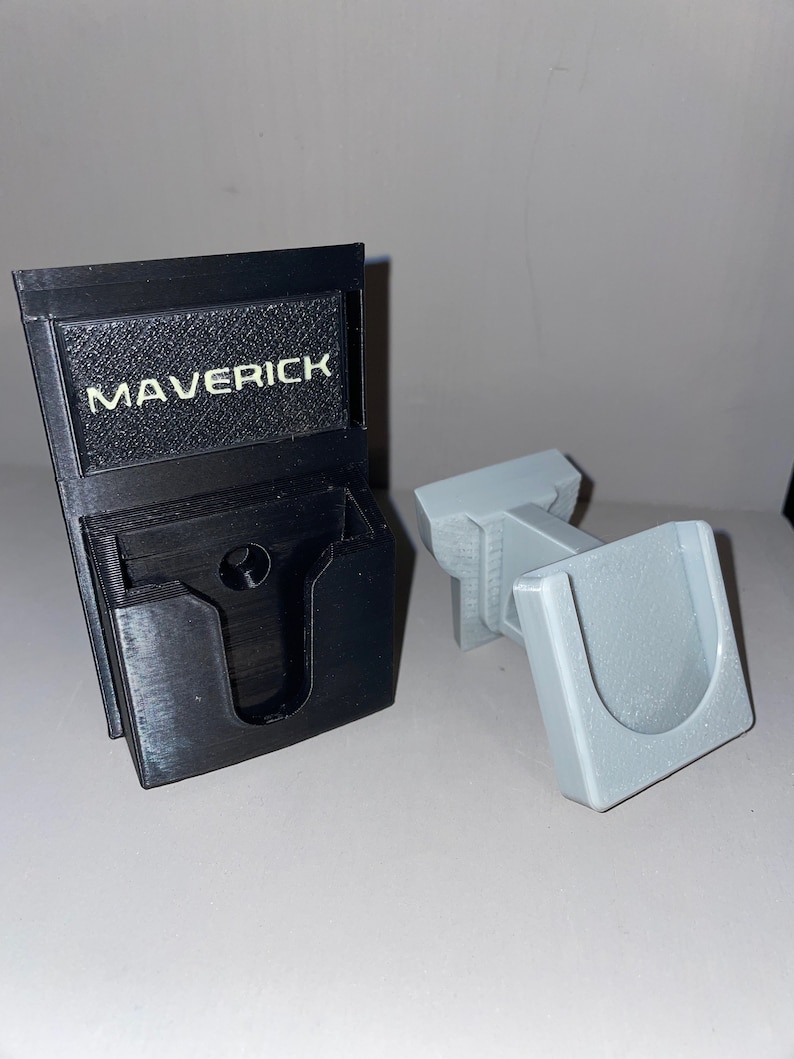Puede incluir: Un soporte de pared de impresi&oacute;n 3D negro con la palabra "MAVERICK" impresa en &eacute;l. El soporte est&aacute; dise&ntilde;ado para sujetar un tel&eacute;fono u otro dispositivo. Tambi&eacute;n se muestra un soporte de tel&eacute;fono de impresi&oacute;n 3D gris.