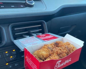 Puede incluir: Una caja roja de Chick-fil-A con un logotipo blanco y el texto "Chick-fil-A" en la parte delantera. La caja contiene una porción de nuggets de pollo. La caja está en el portavasos de un coche.