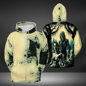 May include: A cream-colored zippered hoodie with a green and black print of a swamp monster and a woman in a swamp setting. The print features the text "Creature from the Black Lagoon".