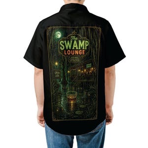 Puede incluir: Camisa negra con botones con un gráfico detallado en la espalda. El gráfico representa una escena de pantano con el texto "The Swamp Lounge" en verde y rojo. El texto adicional incluye "Good Drinks, Strange Company, Low Expectations" y "Open Late".