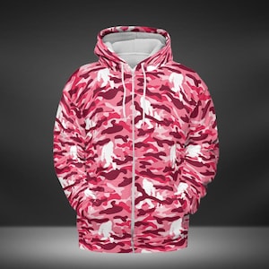 May include: A pink camouflage zip-up hoodie featuring white Bigfoot silhouettes. The hoodie has a white interior lining and a drawstring hood. The camouflage pattern includes shades of pink and burgundy.