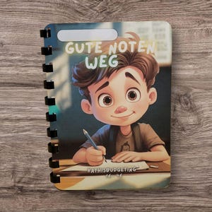 Könnte beinhalten: Ein spiralgebundenes Notizbuch mit einer Cartoon-Illustration eines schreibenden Jungen. Auf dem Cover steht der Text "GUTE NOTEN WEG" und "KATHISBUDGETING". Das Notizbuch hat ein weißes rechteckiges Etikett oben und einen Holzuntergrund.