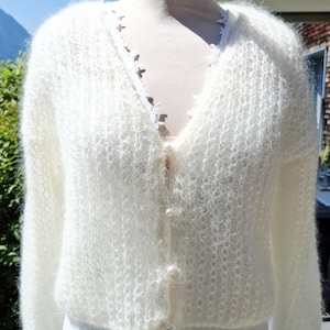 Könnte beinhalten: Eine weiße, flauschige Langarm-Strickjacke mit V-Ausschnitt und einer zarten Spitzenborte am Kragen. Das Kleidungsstück verfügt über eine durchgehende Knopfleiste mit kleinen, runden, perlmuttartigen Knöpfen und eine lockere Strickstruktur.