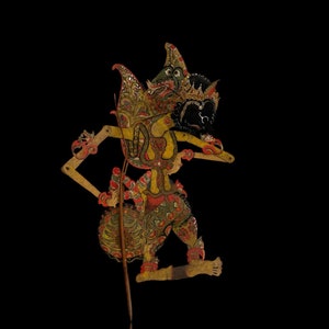 Op de afbeelding: Een traditionele Indonesische Wayang kulit-pop, met ingewikkelde details en levendige kleuren. De pop heeft een gedetailleerde hoofdtooi, een zwart gezicht en is versierd met rode, gele en groene accenten. Gemonteerd op een houten stok, met een reflectie eronder.