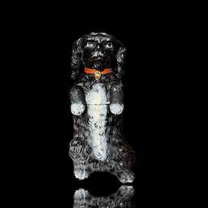 Puede incluir: Figura de perro de cerámica negra con marcas blancas, de pie sobre sus patas traseras con las patas delanteras levantadas. El perro lleva un collar naranja con una campana dorada.