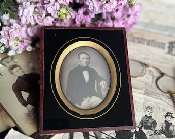 May include: A vintage black and gold framed portrait of a man in a dark suit and white shirt with a bow tie. The portrait is sitting on a table with a stack of old papers and a bouquet of pink and purple flowers.