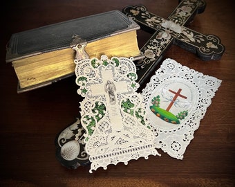 Puede incluir: Tres objetos religiosos vintage: un libro encuadernado en cuero negro, una cruz de encaje blanco con un diseño floral verde y una tarjeta de encaje blanco con una cruz y el texto "Dios es amor" en un círculo.