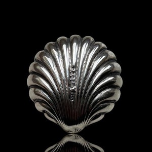 Könnte beinhalten: Eine silberne Zuckerdose in Form einer Muschel mit einem detaillierten, strukturierten Design. Die Schale spiegelt sich auf einer schwarzen Oberfläche.