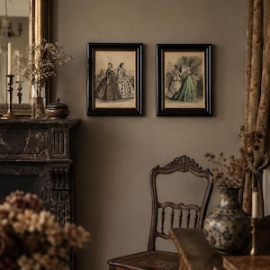 May include: Two rectangular framed prints featuring historical fashion illustrations of figures in long dresses, displayed in black frames with gold inner borders, mounted side-by-side on a solid wall above a wooden chair and fireplace.