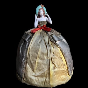 Puede incluir: Una muñeca de porcelana con pelo rojo que lleva un sombrero azul claro y un vestido dorado y gris con un lazo rojo.