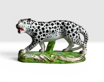 Puede incluir: Figura de leopardo de cerámica con cuerpo blanco y manchas negras. El leopardo está representado caminando sobre una base verde con detalles dorados. La figura tiene la boca roja abierta y ojos verdes.
