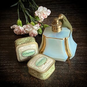 Könnte beinhalten: Eine antike Parfümflasche und zwei Puderdosen. Die Parfümflasche ist hellblau mit goldenen Verzierungen und einem goldenen Zerstäuber. Die Puderdosen sind grün mit dem Wort "Nymphaea". Rosa und weiße Blumen sind im Hintergrund.