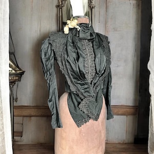 Puede incluir: Una blusa de satén negro con detalles de encaje y un cuello alto. La blusa está en un maniquí y tiene un adorno floral en el cuello.