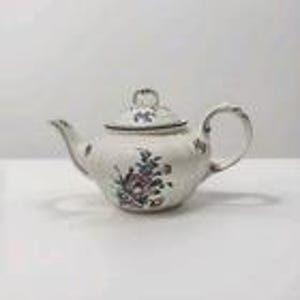 Puede incluir: Una tetera de cerámica blanca con tapa, asa y boquilla. La tetera está decorada con un estampado floral en tonos rosa, azul y verde. La tetera está sobre un fondo blanco.