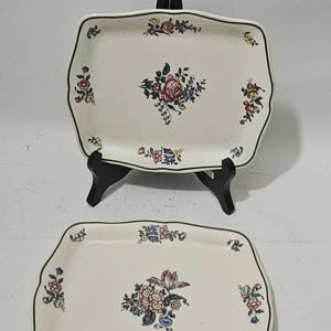 Könnte beinhalten: Zwei rechteckige Servierplatten aus cremeweißer Keramik mit gewelltem Rand und dunkelgrüner Umrandung. Jede Platte ist mit einem zentralen Blumenstrauß und kleineren Blumenmotiven in Rosa, Blau, Gelb und Grün auf der Oberfläche verziert.