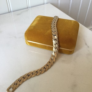 May include: A gold-toned metal bracelet with a textured, linked design rests on a mustard-yellow velvet box. The bracelet's clasp is visible. The box sits on a white marble surface.