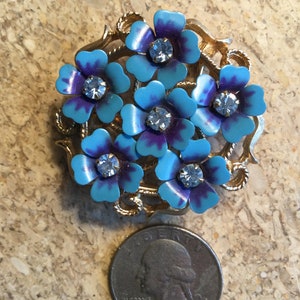 Peut inclure: Une broche dorée avec un groupe de fleurs en émail bleu et violet avec des centres de strass clairs.