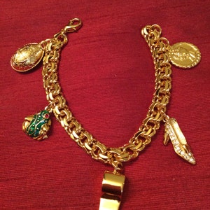 May include: A gold charm bracelet with a variety of charms, including a whistle, a frog, a shoe, and a coin. The bracelet is made of a thick, gold chain.