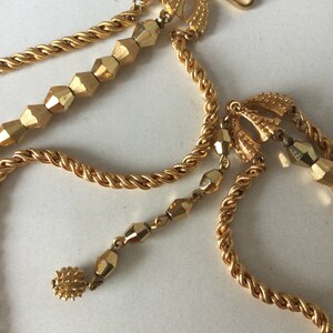 Può includere: Una collana dorata con una catena intrecciata e perline geometriche. La collana ha due chiusure e una perla pendente.