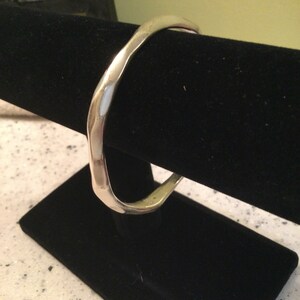 May include: A silver bangle bracelet with a hammered finish. The bracelet is displayed on a black velvet stand.