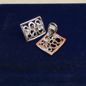May include: A pair of silver clip-on earrings with a geometric design. The earrings are square shaped and feature a delicate, intricate pattern.