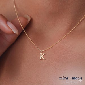 May include: A delicate gold necklace with a small pendant in the shape of the letter "K". The pendant is suspended from a fine chain. The necklace is shown on a person's neck, with the brand name "mira moon" at the bottom.