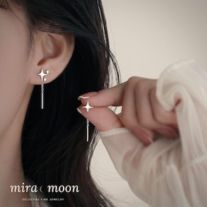 Op de afbeelding: Zilveren ster-vormige oorbellen met een bungelende staaf. Eén oorbel heeft een cluster van sterren, terwijl de andere door een hand wordt vastgehouden. De oorbellen maken deel uit van de "mira moon" Celestial Fine Jewelry collectie.