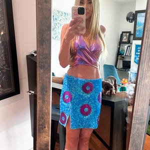 May include: A holographic wrap top and a sequined mini skirt. The top has a V-neck design. The skirt is blue with large, pink sequined circles. The skirt has a slit on the left side.