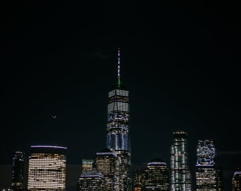 May include: A night view of the New York City skyline with the Freedom Tower lit up and a full moon in the dark sky.