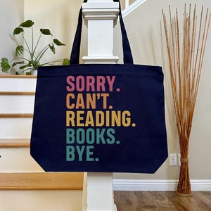 May include: A navy blue canvas tote bag with the text "SORRY. CAN'T. READING. BOOKS. BYE." in a rainbow color gradient.