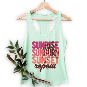 May include: A light green tank top with a white racerback design. The front of the tank top has the text "Sunrise Sunburn Sunset Repeat" in pink, red, and brown with a palm tree graphic.