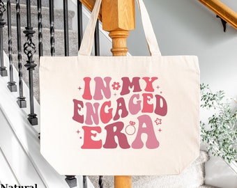 May include: A natural canvas tote bag with the text "IN MY ENGAGED ERA" in pink and red retro font with a diamond ring graphic.