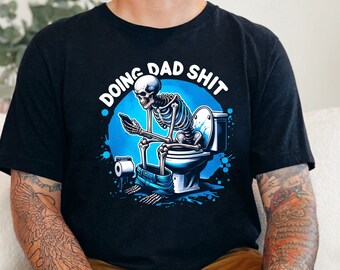 May include: Black t-shirt with the text "DOING DAD SHIT" above a cartoon skeleton sitting on a toilet, looking at a phone. The design features blue and white accents.