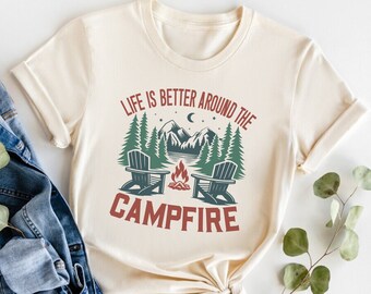 May include: Sand-colored t-shirt with the text "LIFE IS BETTER AROUND THE CAMPFIRE." The design features a campfire, two chairs, trees, mountains, and a crescent moon. A denim jacket is draped on the side.