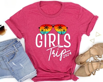 May include: A heather raspberry t-shirt with the words "GIRLS TRIP 2024" in white script. The shirt features a graphic of sunglasses with a sunset and palm trees. The shirt is paired with denim shorts and sandals.
