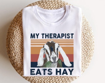 Peut inclure: T-shirt blanc avec un graphique vintage d'une chèvre mangeant du foin. Le texte "My Therapist Eats Hay" est imprimé sur le t-shirt.