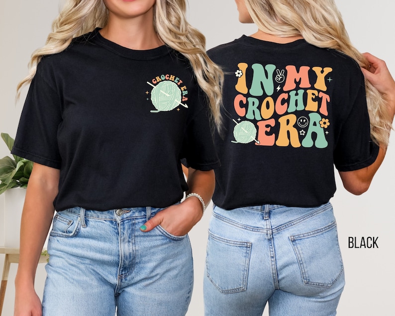 K&ouml;nnte beinhalten: Schwarzes T-Shirt mit dem Text "CROCHET ERA" und einer Grafik aus Garn und einem H&auml;kelhaken auf der Vorderseite. Auf der R&uuml;ckseite des Shirts steht der Text "IN MY CROCHET ERA" in farbenfrohen Retro-Lettern sowie zus&auml;tzliche Grafiken.