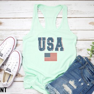 K&ouml;nnte beinhalten: Ein mintgr&uuml;nes Tanktop mit einem verwaschenen blauen "USA"-Motiv und einer kleinen amerikanischen Flagge. Das Tanktop liegt auf einem wei&szlig;en Holzgrund.
