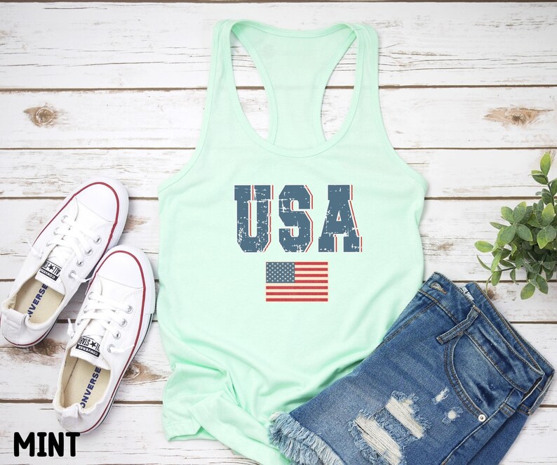 K&ouml;nnte beinhalten: Ein mintgr&uuml;nes Tanktop mit einem verwaschenen blauen "USA"-Motiv und einer kleinen amerikanischen Flagge. Das Tanktop liegt auf einem wei&szlig;en Holzgrund.