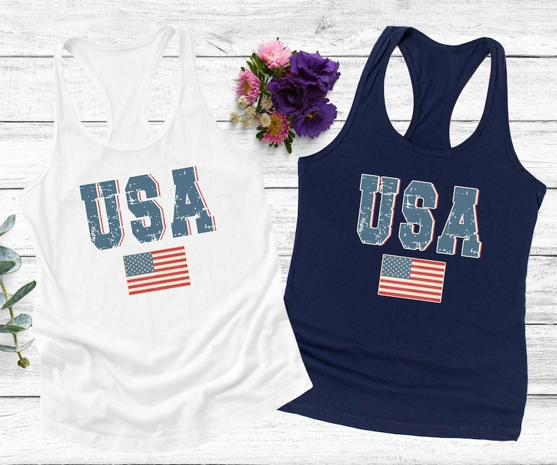 K&ouml;nnte beinhalten: Zwei Tanktops mit dem Wort "USA" in blau bedruckt, mit einem abgenutzten Look und einer kleinen amerikanischen Flagge darunter. Das Tanktop links ist wei&szlig; und das Tanktop rechts ist marineblau.
