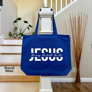 May include: Royal blue tote bag with white text that reads "JESUS the way the truth the life". The bag is hanging on a white post. The background includes a staircase, plants, and wall decor.