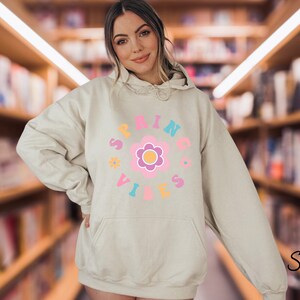 Op de afbeelding: Een licht beige hoodie met een grafisch ontwerp van een roze bloem met een gele kern en de tekst "SPRING VIBES" in een cirkel rond de bloem.
