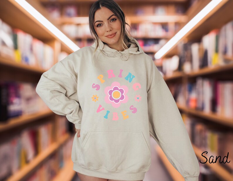 Op de afbeelding: Een licht beige hoodie met een grafisch ontwerp van een roze bloem met een gele kern en de tekst "SPRING VIBES" in een cirkel rond de bloem.