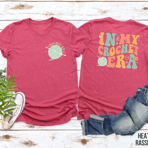 K&ouml;nnte beinhalten: Heather-Himbeerfarbenes T-Shirt mit dem Text "CROCHET ERA" und einer Garnkn&auml;uel-Grafik auf der Vorderseite. Auf der R&uuml;ckseite des Shirts steht "IN MY CROCHET ERA" mit Peace-Zeichen, Blumen und einem Smiley. Eine Jeans ist daneben gefaltet.
