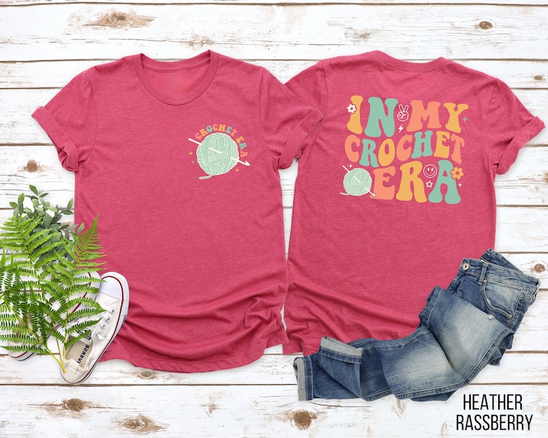 K&ouml;nnte beinhalten: Heather-Himbeerfarbenes T-Shirt mit dem Text "CROCHET ERA" und einer Garnkn&auml;uel-Grafik auf der Vorderseite. Auf der R&uuml;ckseite des Shirts steht "IN MY CROCHET ERA" mit Peace-Zeichen, Blumen und einem Smiley. Eine Jeans ist daneben gefaltet.